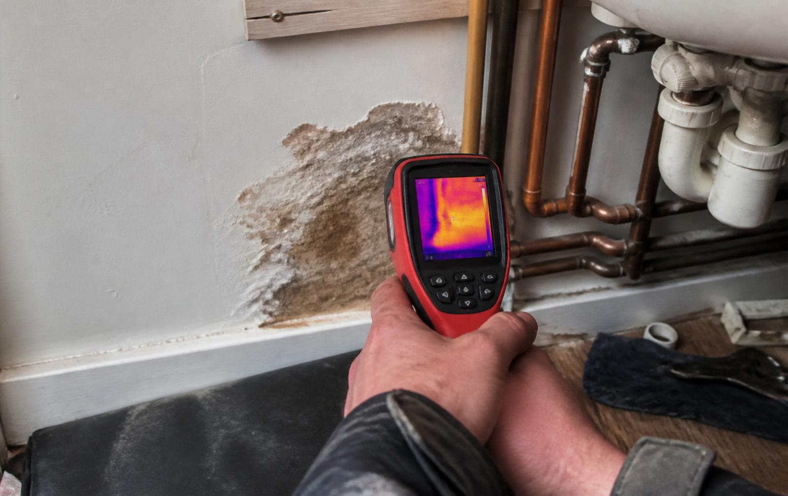 Handheld thermal imaging camera beside exposed copper pipework in a damp wall — Ash Villa leak detection and repair in West London
