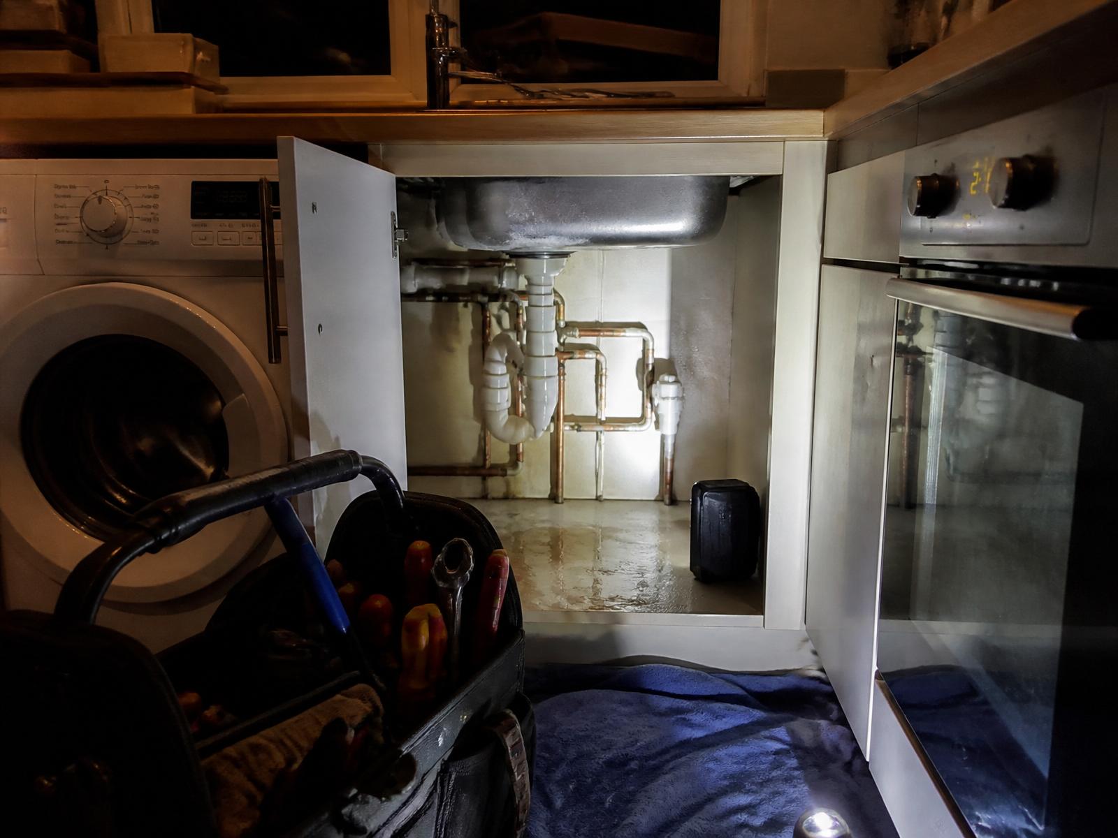 Emergency plumber's tool bag and work light beside an isolated burst pipe under a kitchen sink at night — Ash Villa 24/7 emergency plumbing in West London